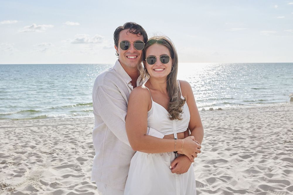 Happy engaged couple in white outfits embracing on a sunny tropical beach.