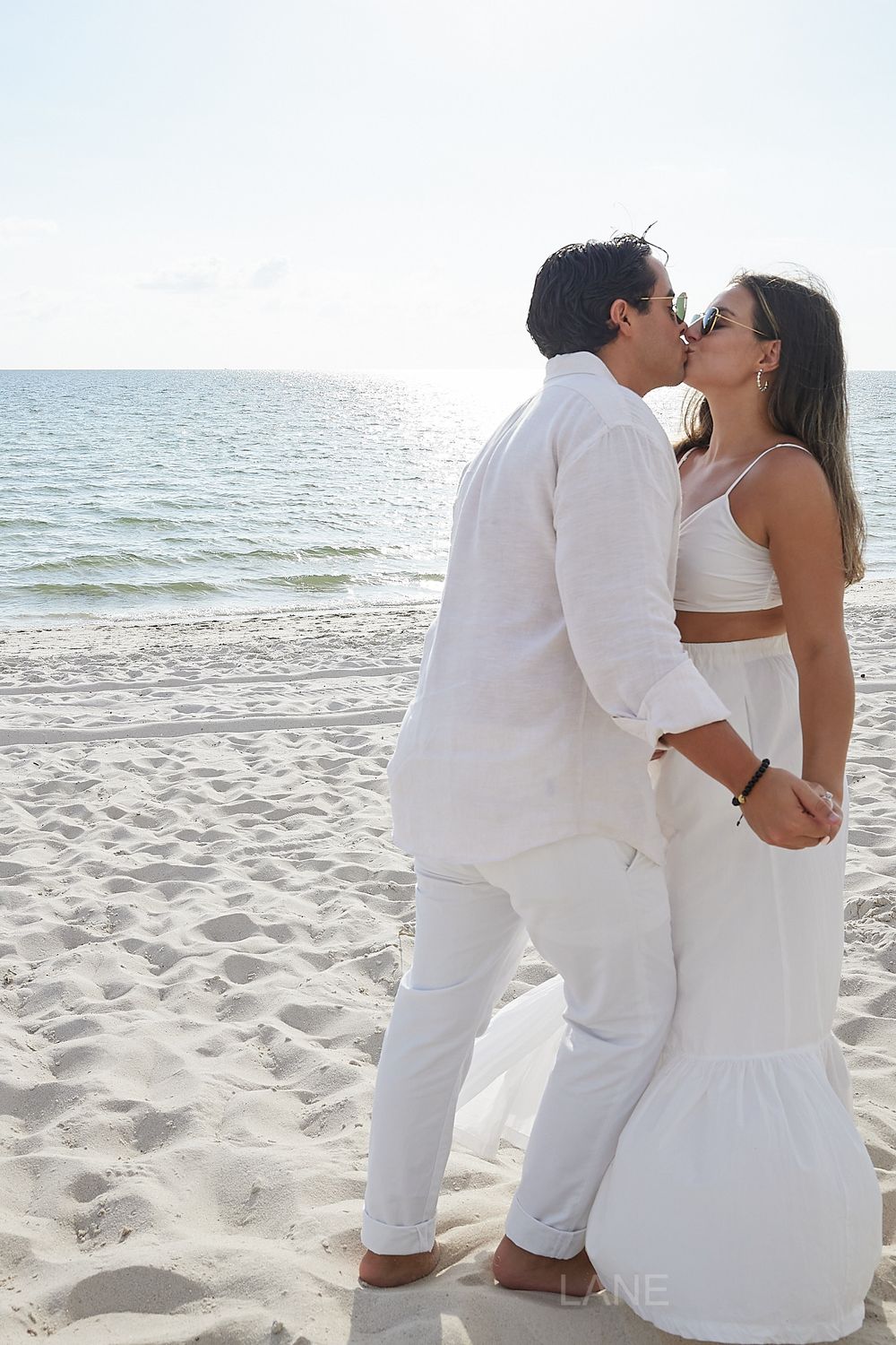 Couple sharing a romantic kiss on a white sandy beach with ocean backdrop.