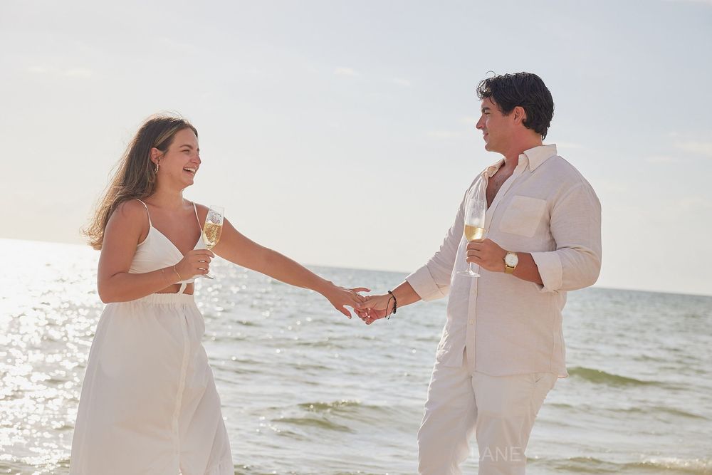 Happy couple in white holding hands and champagne glasses on a sunny beach.