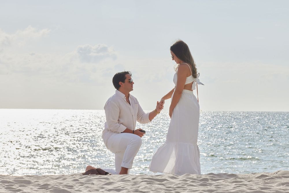 Man proposing on one knee to woman in white dress on a sunny beach by the ocean.