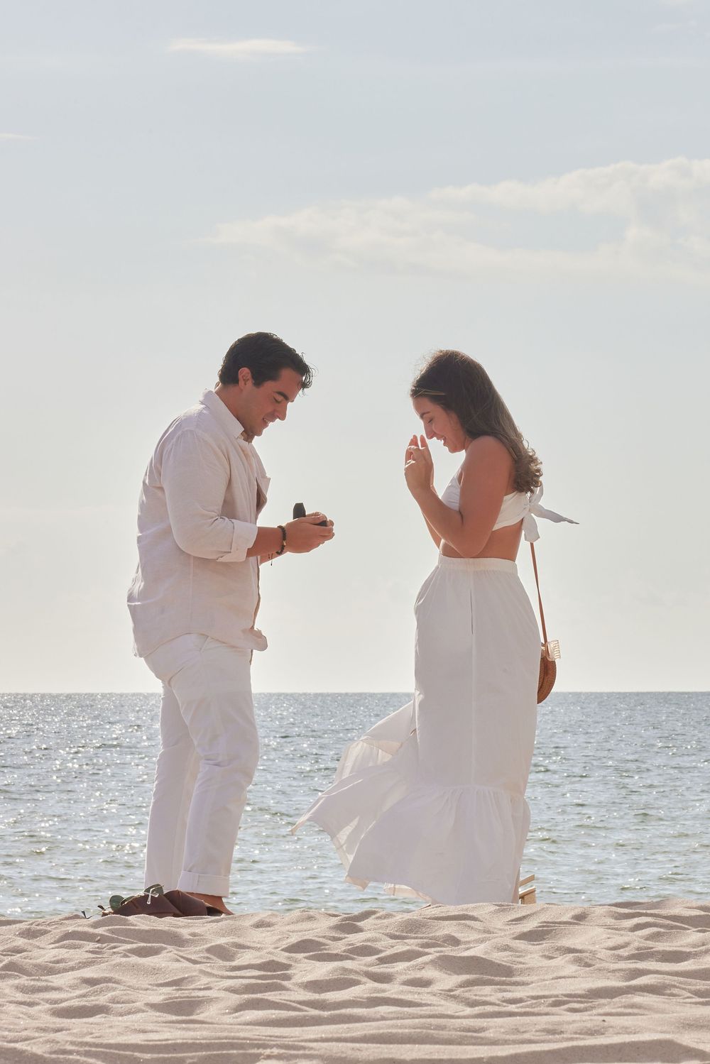 Man presenting engagement ring to emotional woman on a sandy beach at sunset.