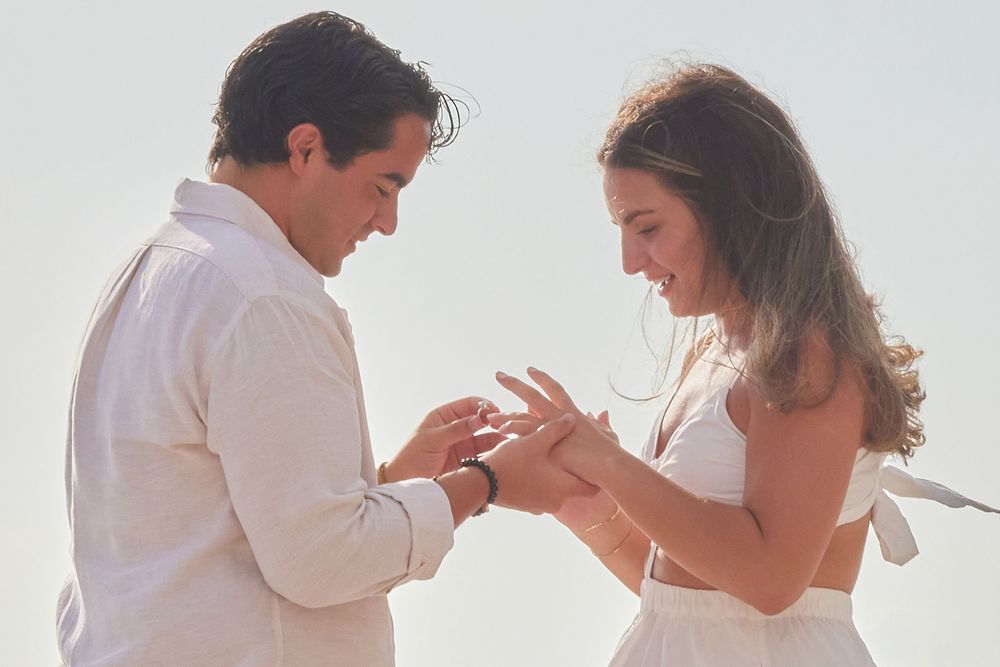 Couple exchanging rings on a sunlit beach, smiling joyfully at each other.