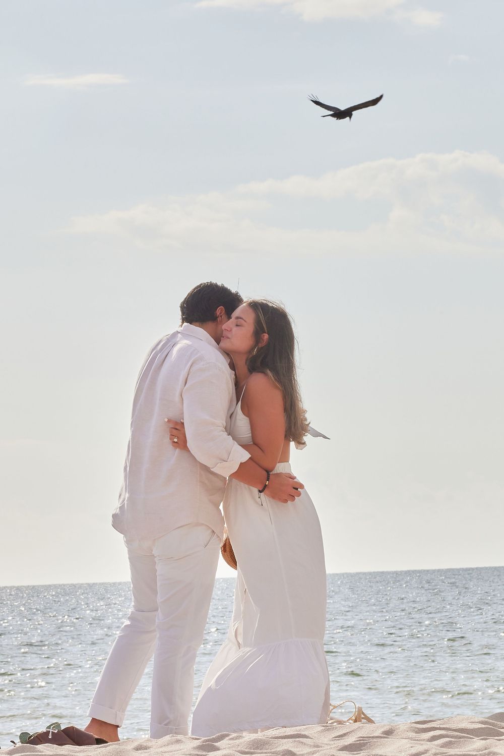 Newly engaged couple sharing a kiss on the beach with a bird flying overhead.