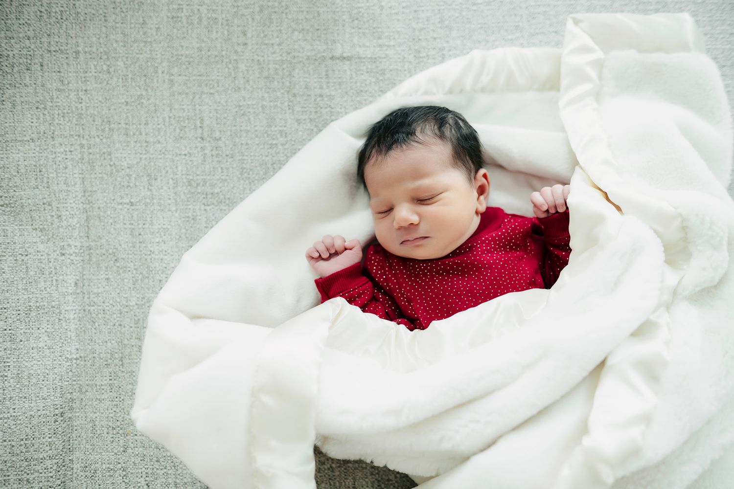 Newborn baby sleeping peacefully wrapped in a red outfit and white blanket on a gray background.