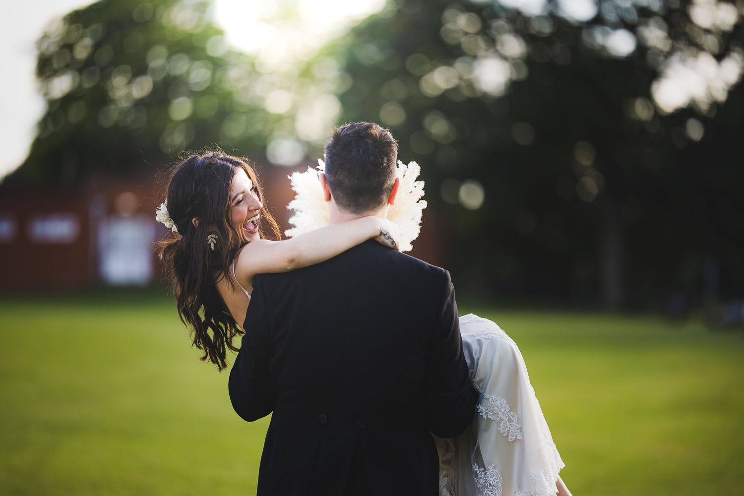 A bride and groom embrace outdoors on a lush green lawn, with the bride smiling over the groom's shoulder on their wedding day.