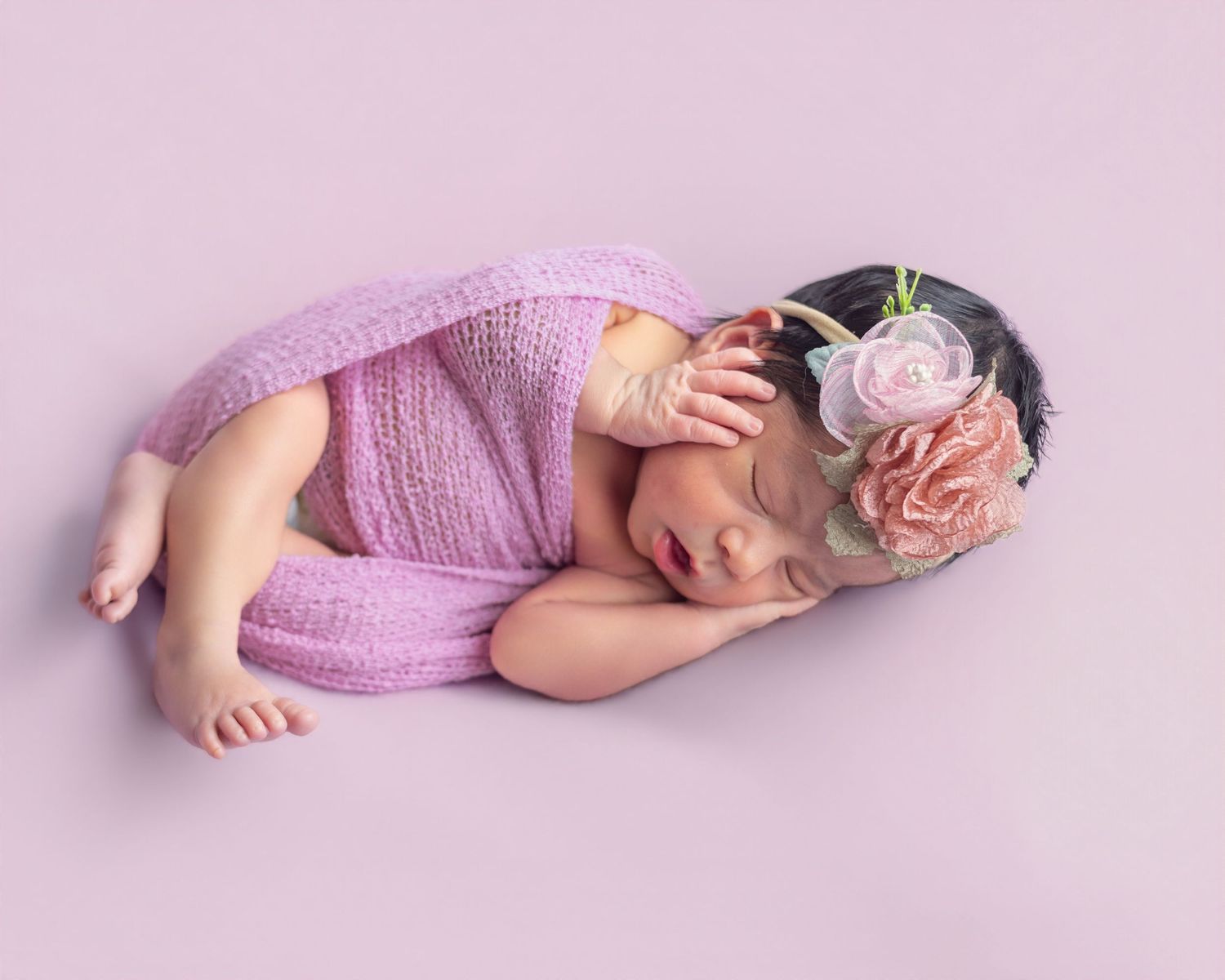 Sleeping newborn baby girl in pink knit wrap and floral headband on lavender background.