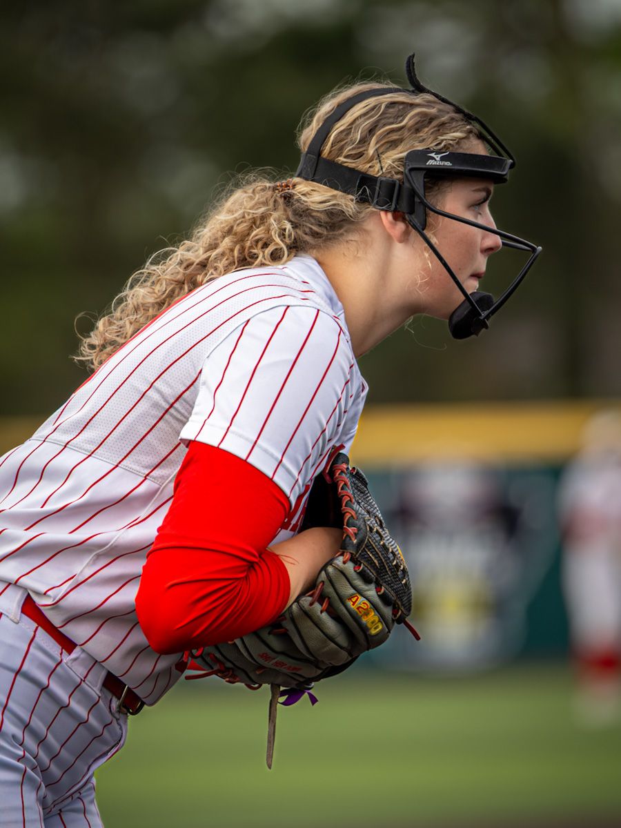 Female softball pitcher in white pinstripe uniform with face guard holds glove ready on the mound.