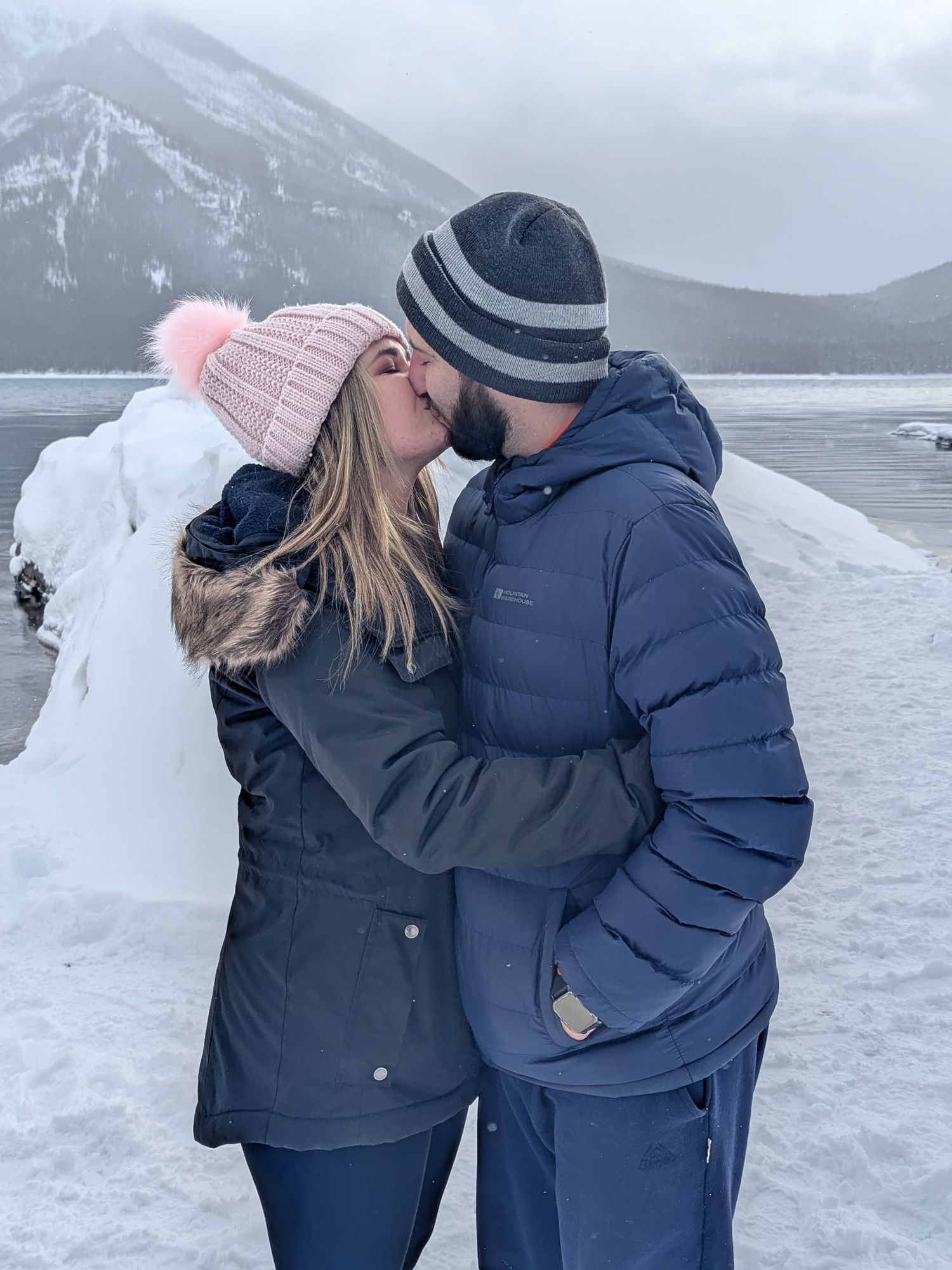 Couple kissing in winter landscape near snowy mountains, wearing navy blue jackets and knit hats.