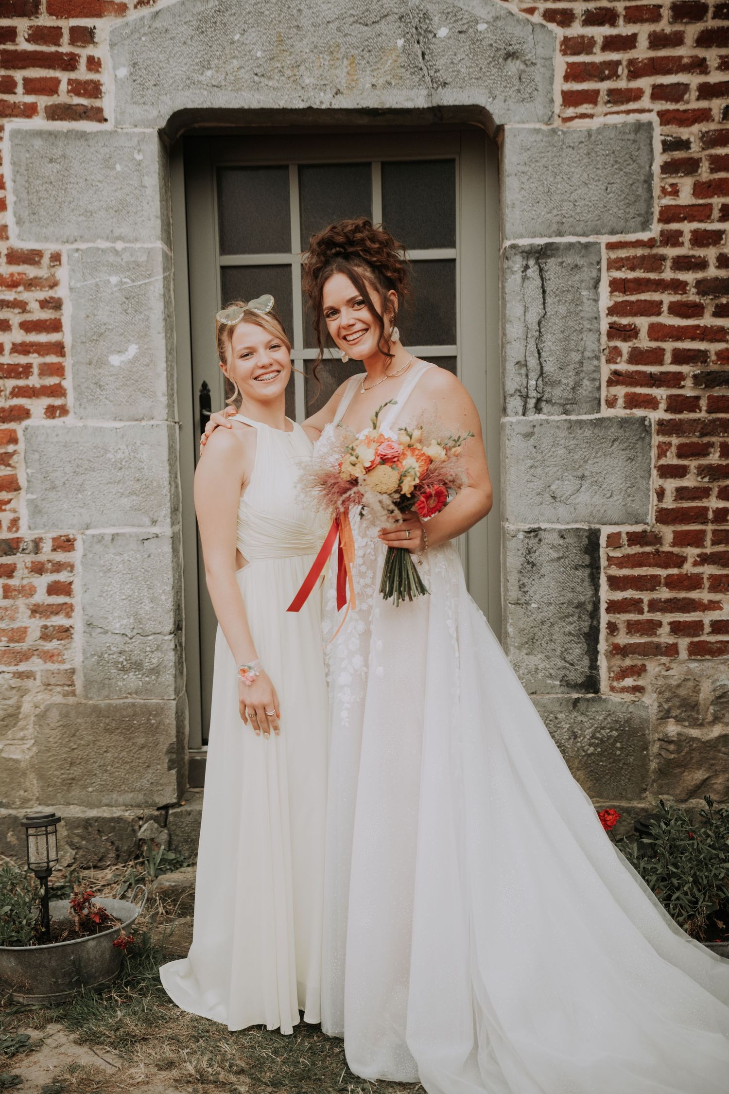Two brides in white wedding gowns pose together holding a colorful bouquet in front of a rustic brick building.