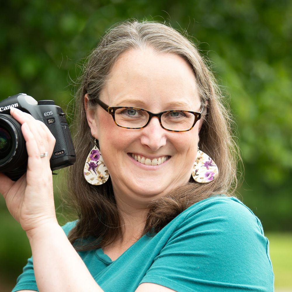 Smiling woman with glasses holding a Canon camera outdoors, wearing teal shirt and floral earrings.