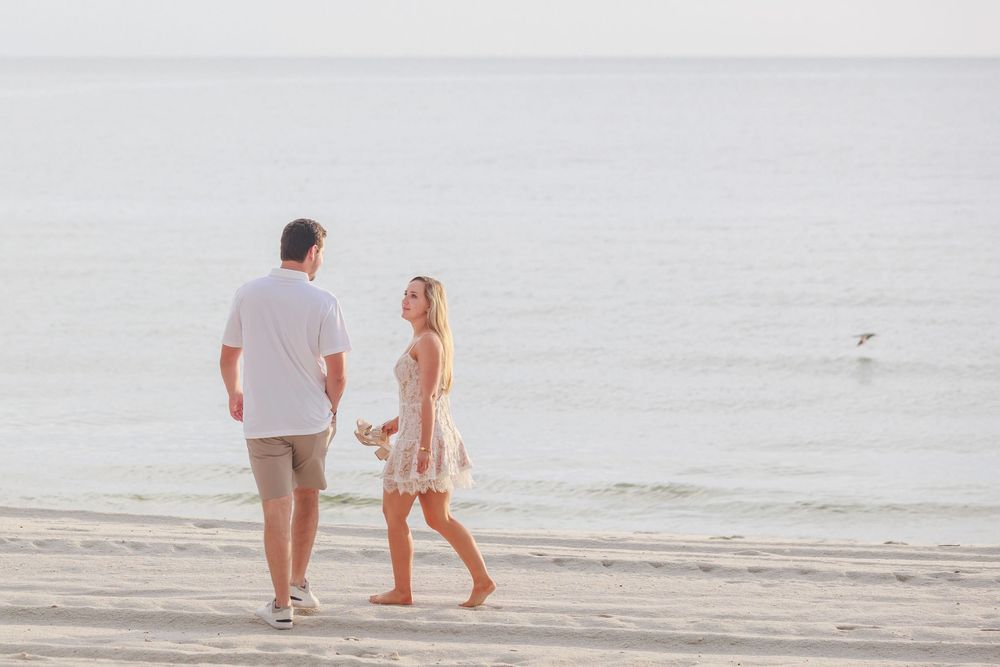Couple walking together along a peaceful beach shoreline, man in white shirt holding flowers.