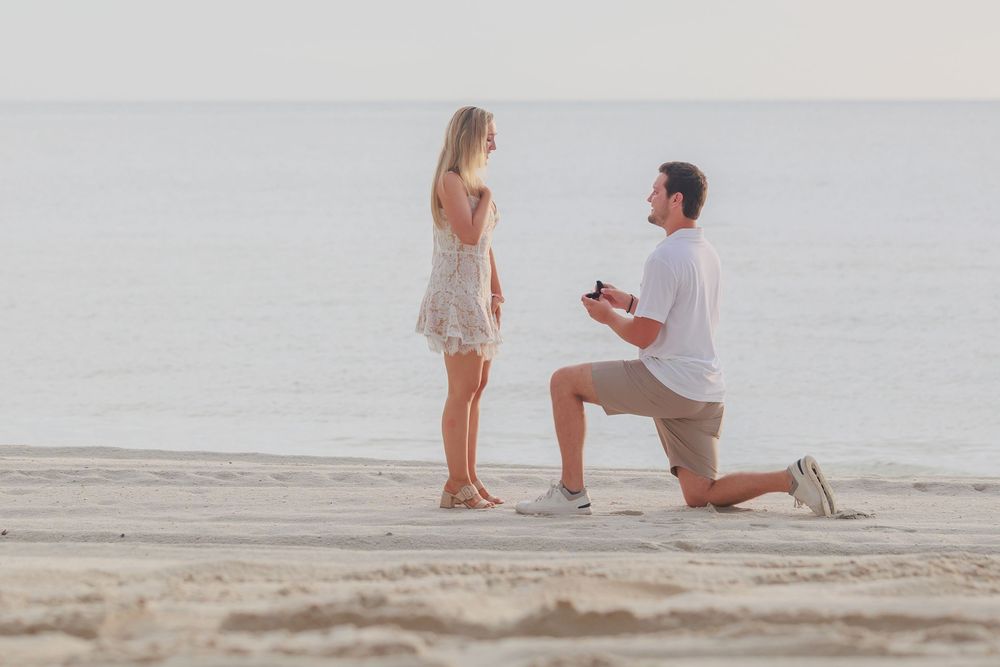 Man on one knee proposing to woman on a serene beach at sunset, holding open ring box.