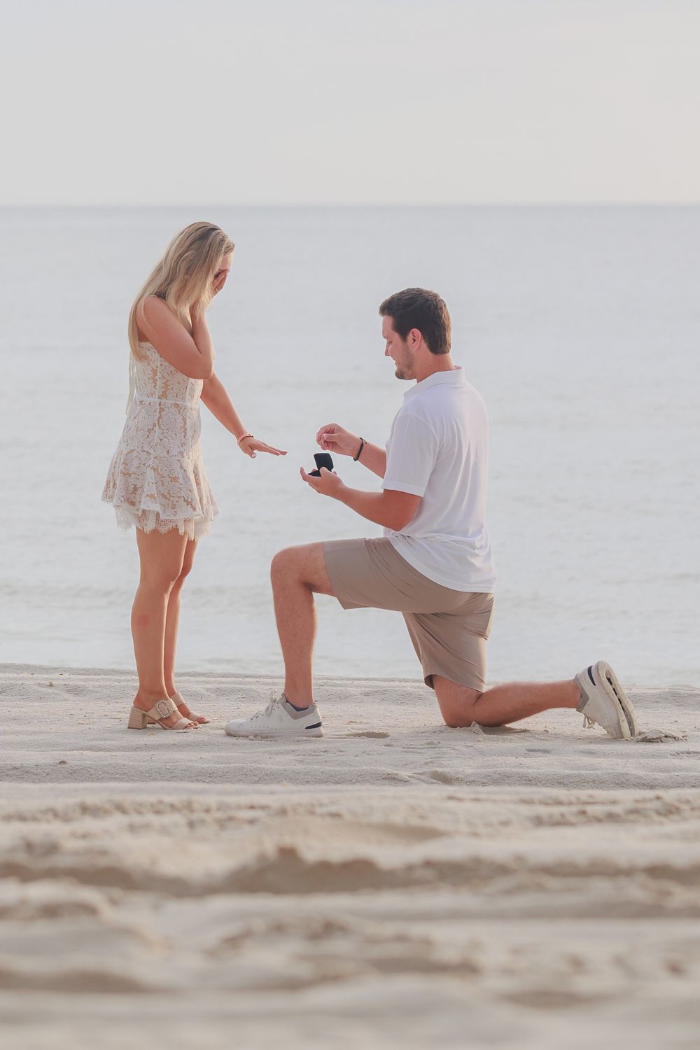 Man proposing on one knee to woman on a sunny beach, presenting an open ring box.