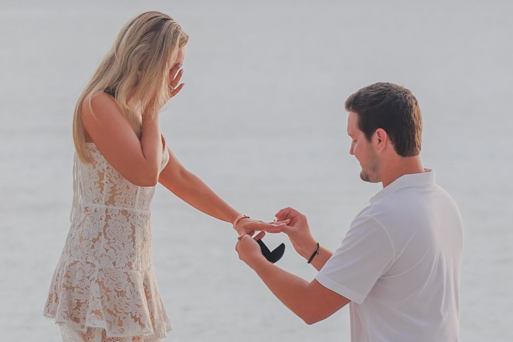 Man kneeling on beach placing engagement ring on womans finger during a romantic seaside proposal.