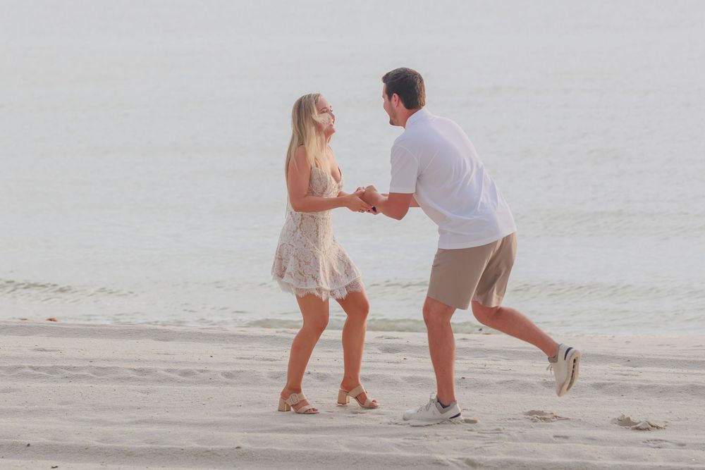 Joyful couple dancing and holding hands on a sandy beach with ocean waves in the background.
