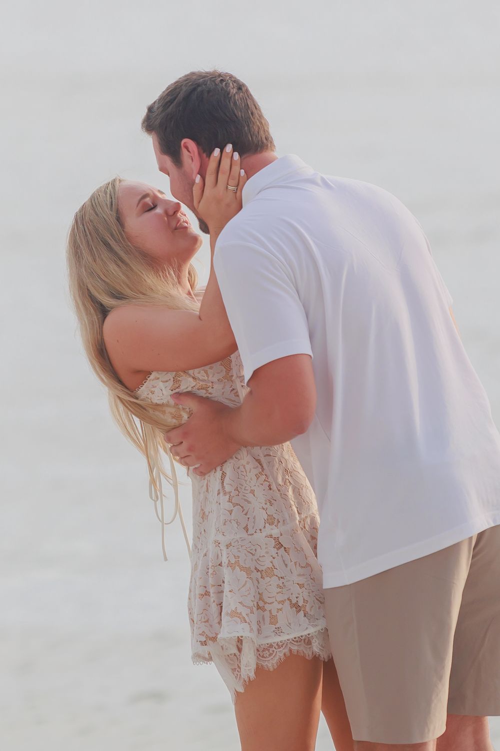 Newly engaged couple sharing a romantic kiss on the beach, woman in lace dress touching mans face.