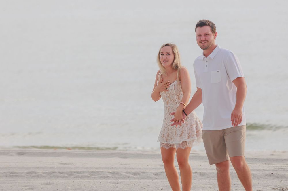 Happy couple walking hand in hand on a misty beach, woman in lace dress admiring her engagement ring.