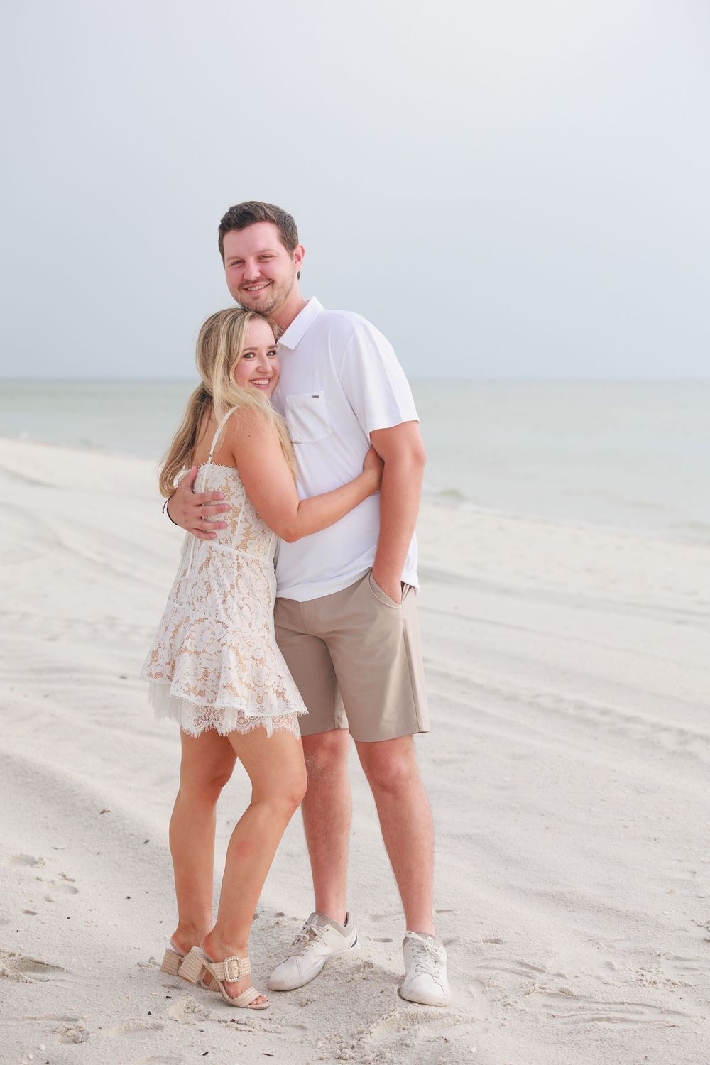 Smiling couple embracing on a white sandy beach, man in white polo and khaki shorts with blonde woman in lace dress.