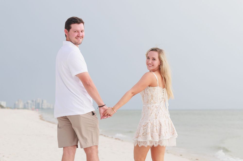 Couple holding hands on a sandy beach, smiling and looking back at the camera near the ocean shoreline.