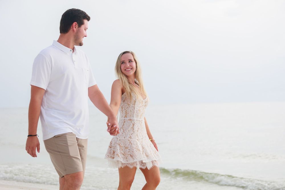Happy couple walking hand in hand along the shoreline smiling at each other on the beach.