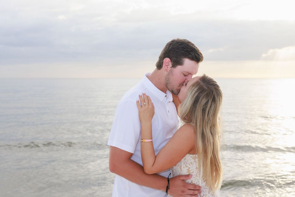 Couple sharing a tender kiss on the beach with calm ocean waves in the background.