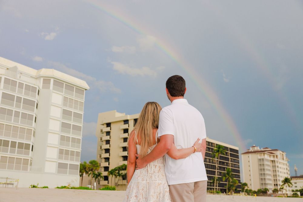 Engaged couple embracing while watching a beautiful double rainbow over beachfront buildings.