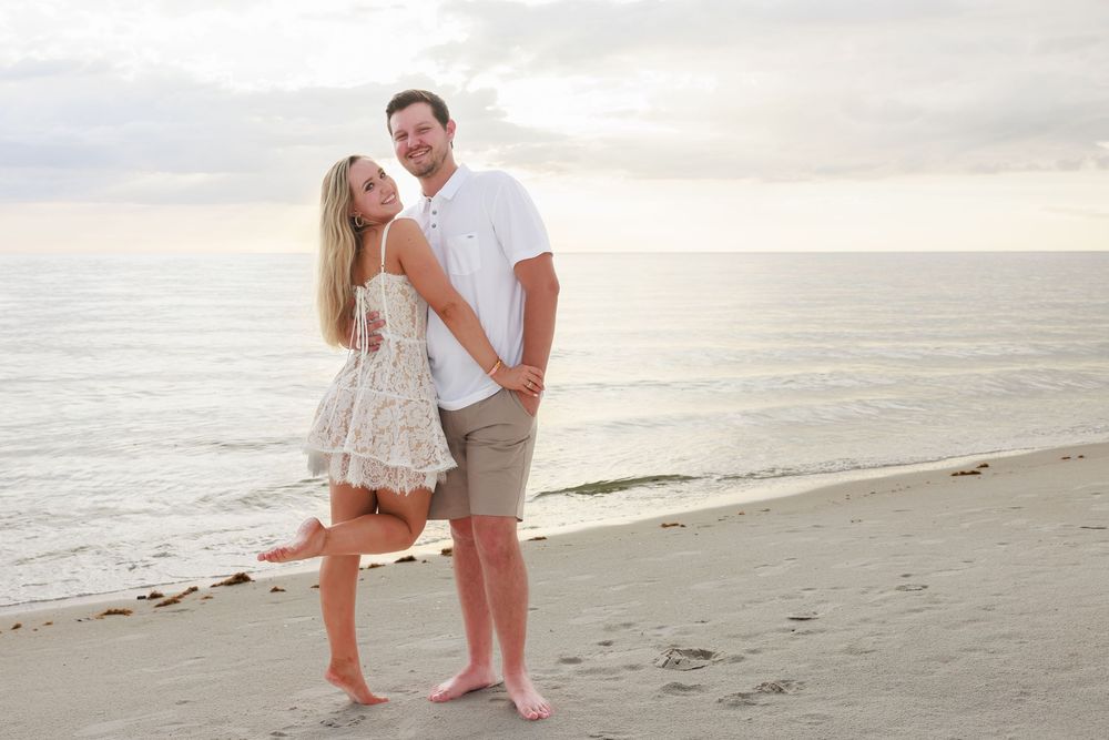 Newly engaged couple holding hands and smiling on a sandy beach at golden hour.