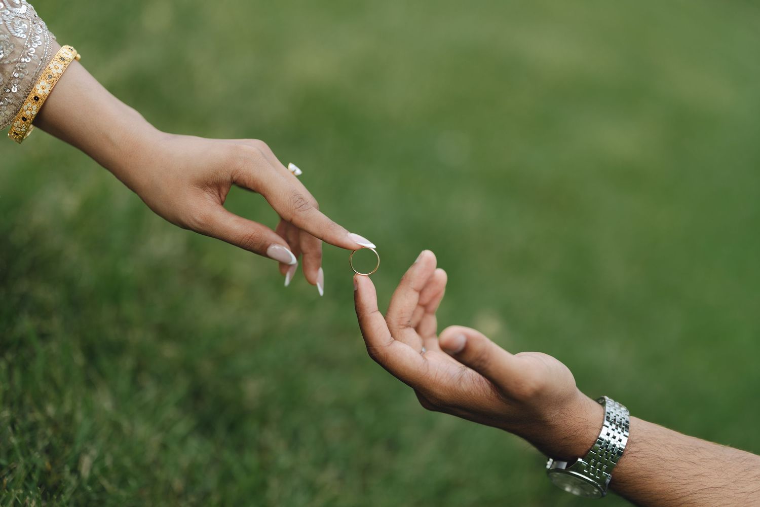 Two hands reaching toward each other outdoors, one with gold bracelet and manicured nails, other with silver watch.