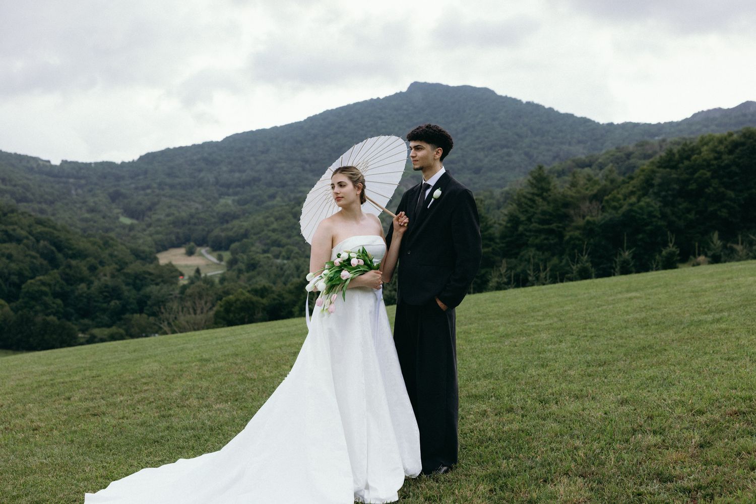 Bride and groom stand on green hillside, bride in white gown holding bouquet and parasol, mountains in background.