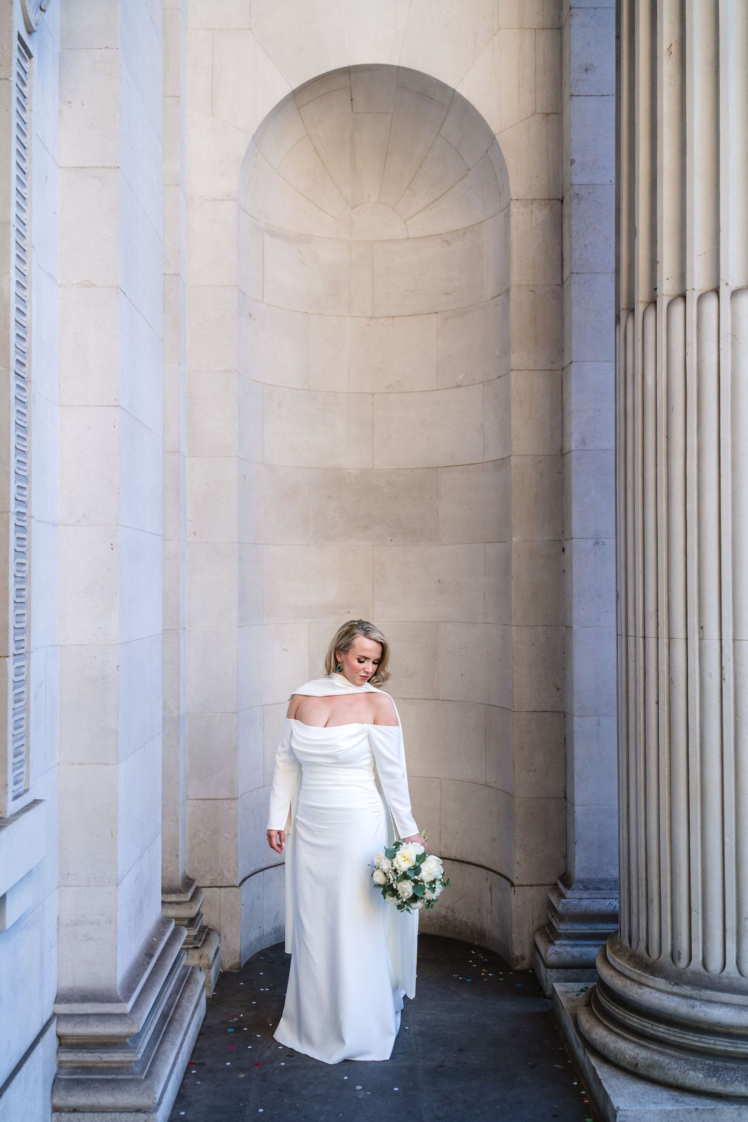 Bride in white off-shoulder gown holding bouquet stands between stone columns and arched alcove.