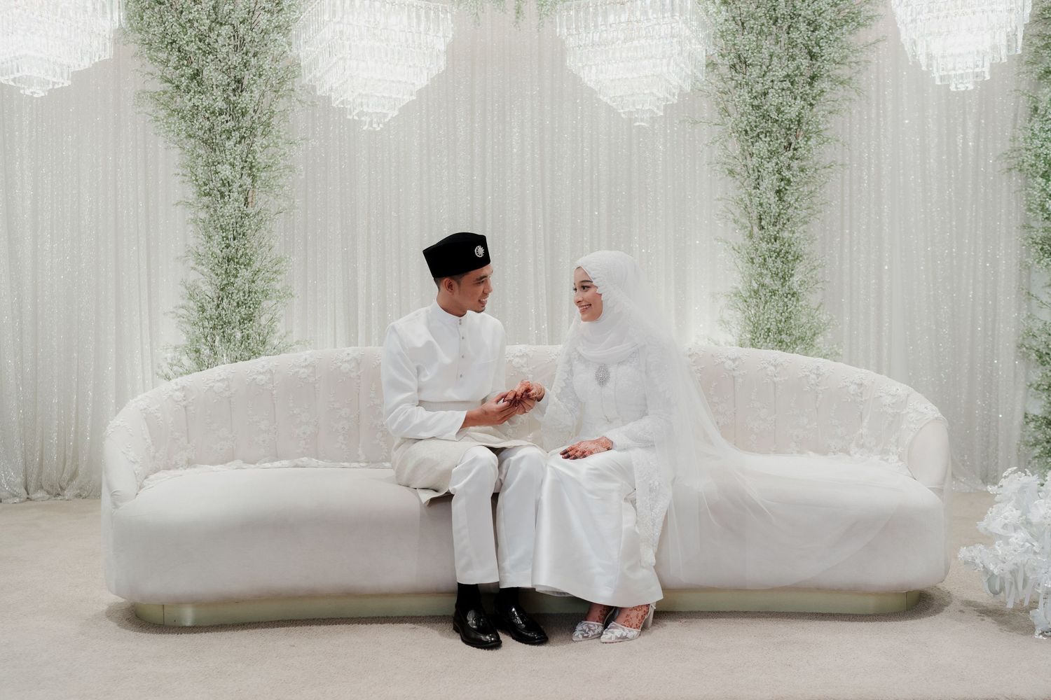 Muslim couple in traditional Javanese wedding attire sitting together at an outdoor ceremony with floral decorations and ocean view.