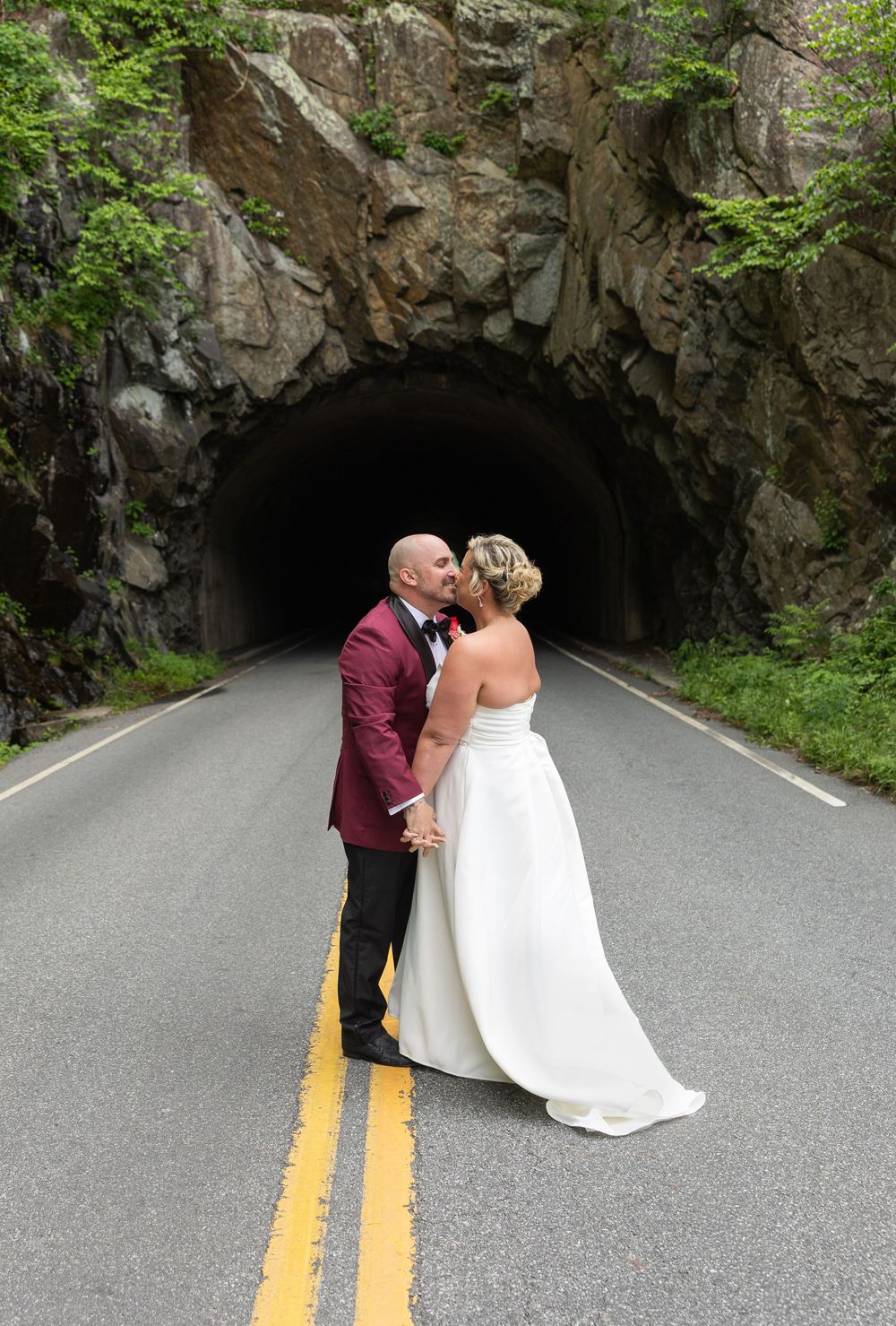 Bride and groom kissing on a road in front of a rock tunnel, groom in burgundy suit, bride in white gown.