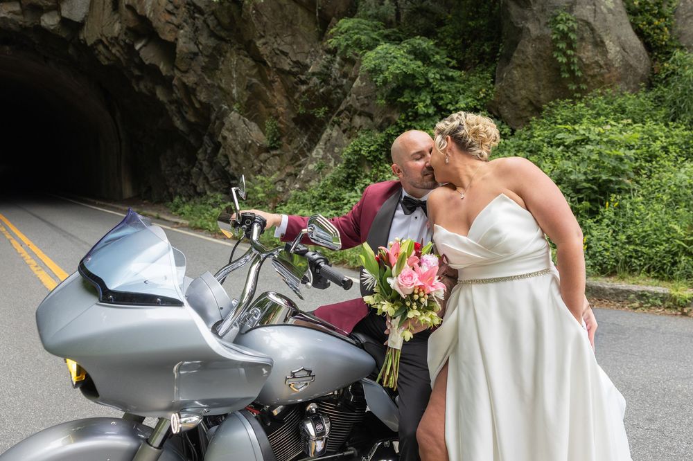 Bride and groom sharing a kiss beside a silver Harley-Davidson motorcycle near a tunnel on their wedding day.