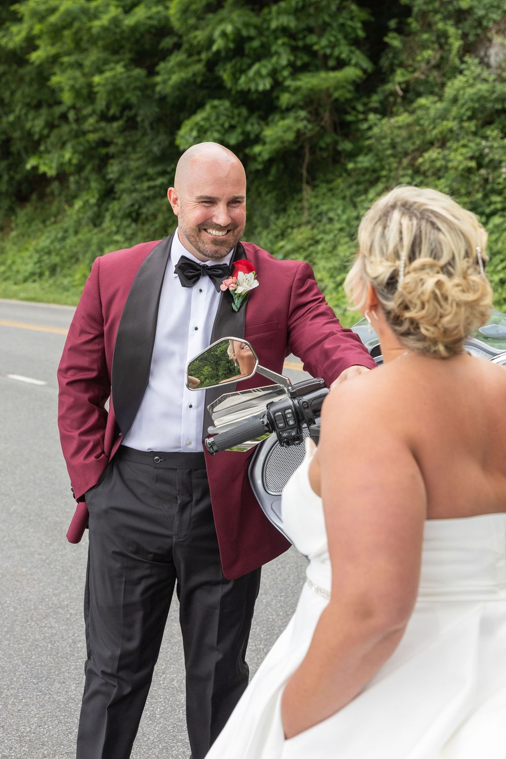 Smiling bald groom in burgundy tuxedo shares a joyful first look moment with his bride on a scenic road.