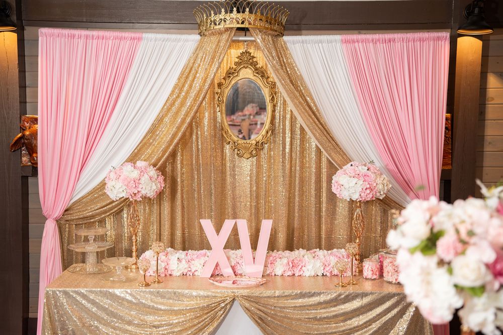 Royal quinceañera backdrop with gold crown, pink drapes, floral arrangements, and XV letters on sequin table.