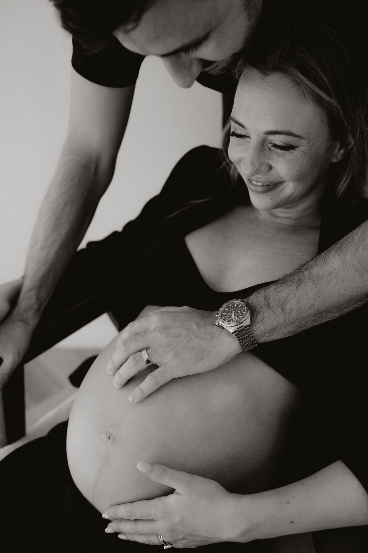 Black and white maternity photo of a smiling pregnant woman and partner tenderly cradling her baby bump together.