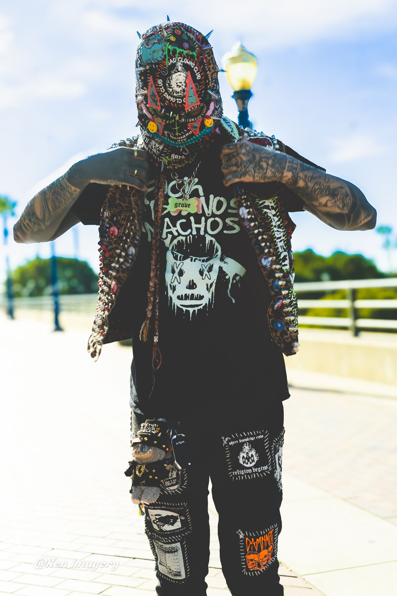 Person in elaborate punk costume with spiked mask, patched jacket, and graphic tee posing outdoors on a sunny day.