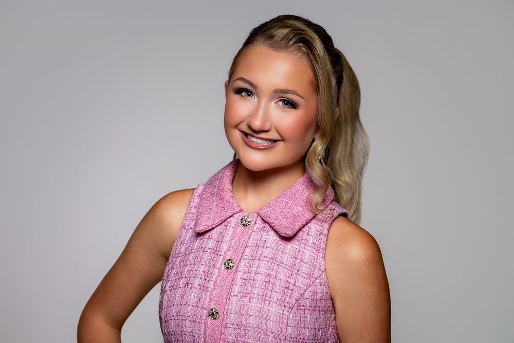 Smiling young woman with blonde hair in a pink tweed sleeveless dress poses against a gray background.