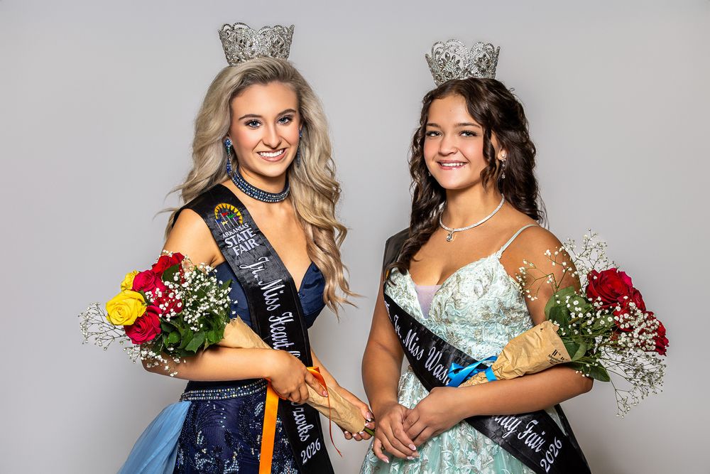 Two young women in formal gowns, crowns, and sashes holding bouquets pose together at a beauty pageant.