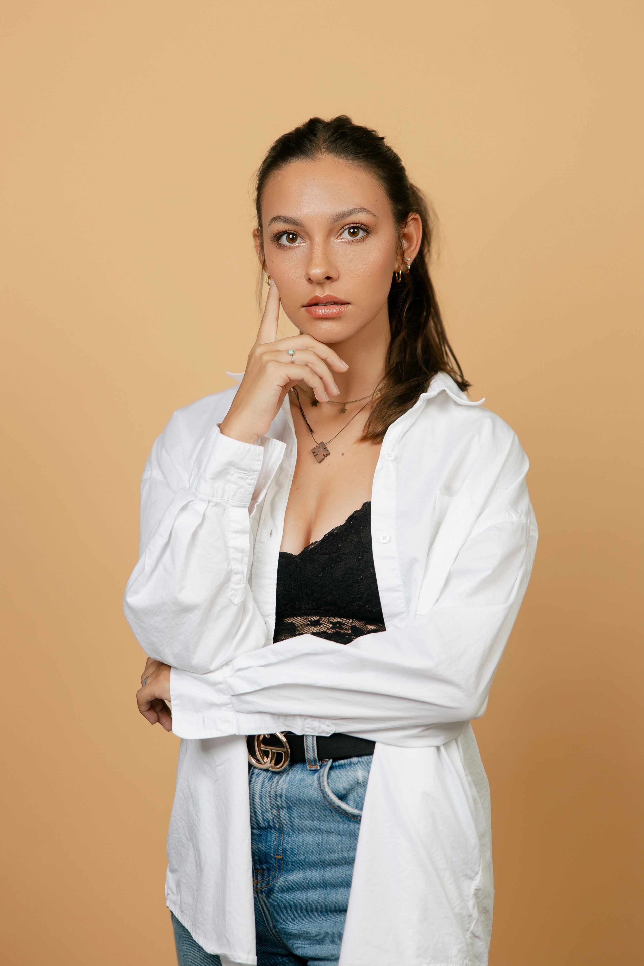 Young woman in white oversized shirt, black lace top, jeans, and Gucci belt posing thoughtfully on beige background.