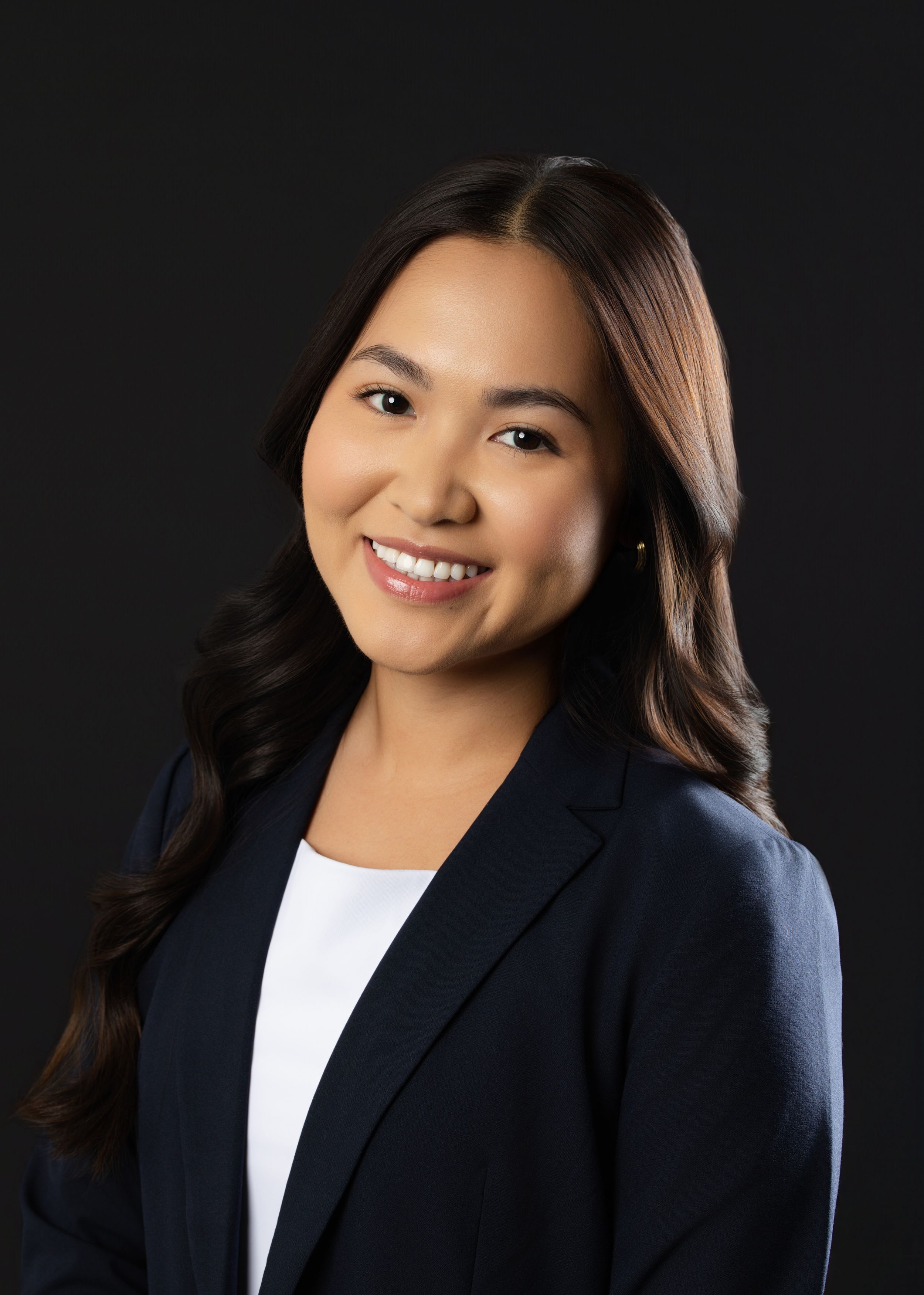 Professional headshot of a smiling young woman in a navy blazer and white top against a dark background.