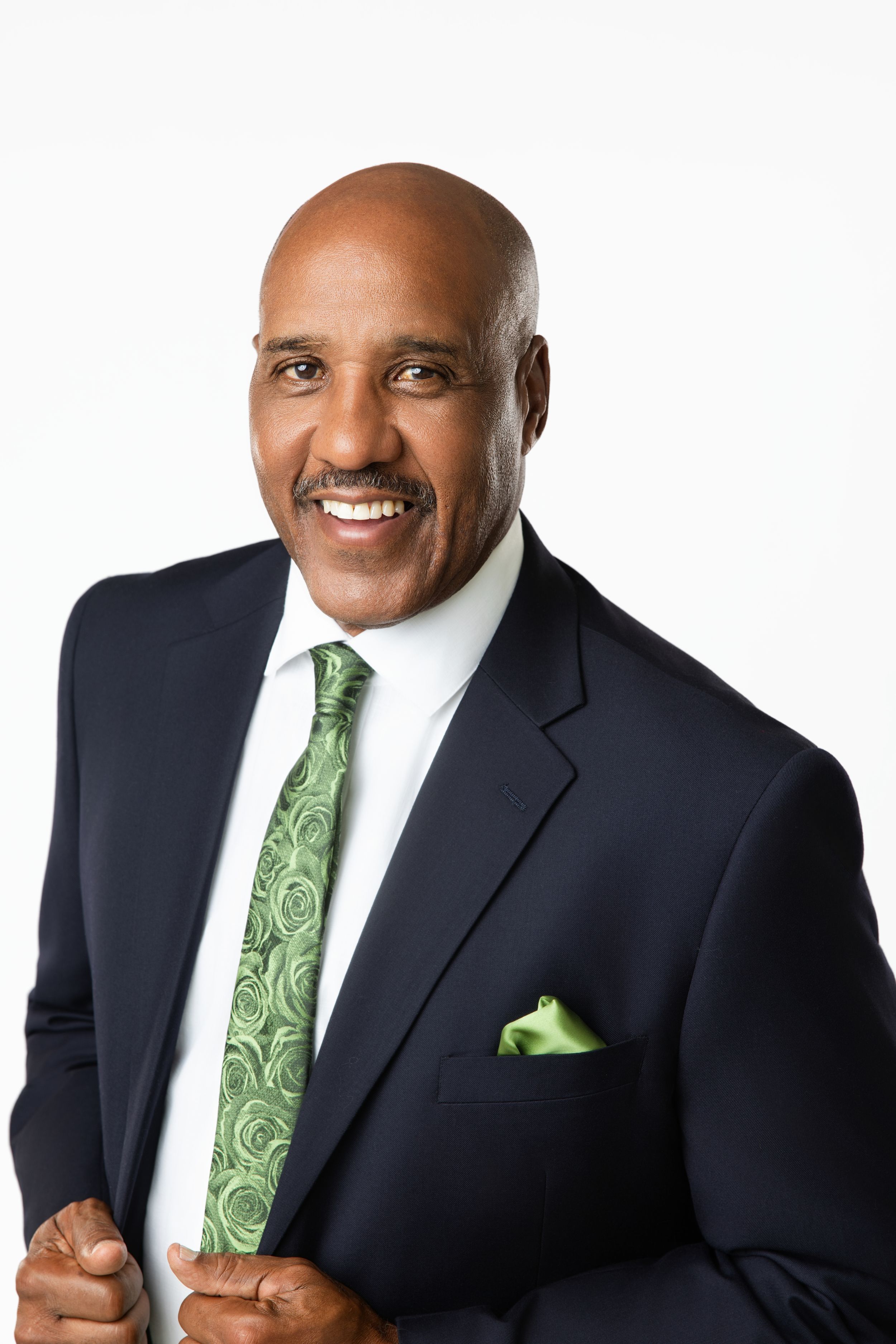 Smiling bald businessman wearing a navy suit with green tie and pocket square poses confidently against white background.