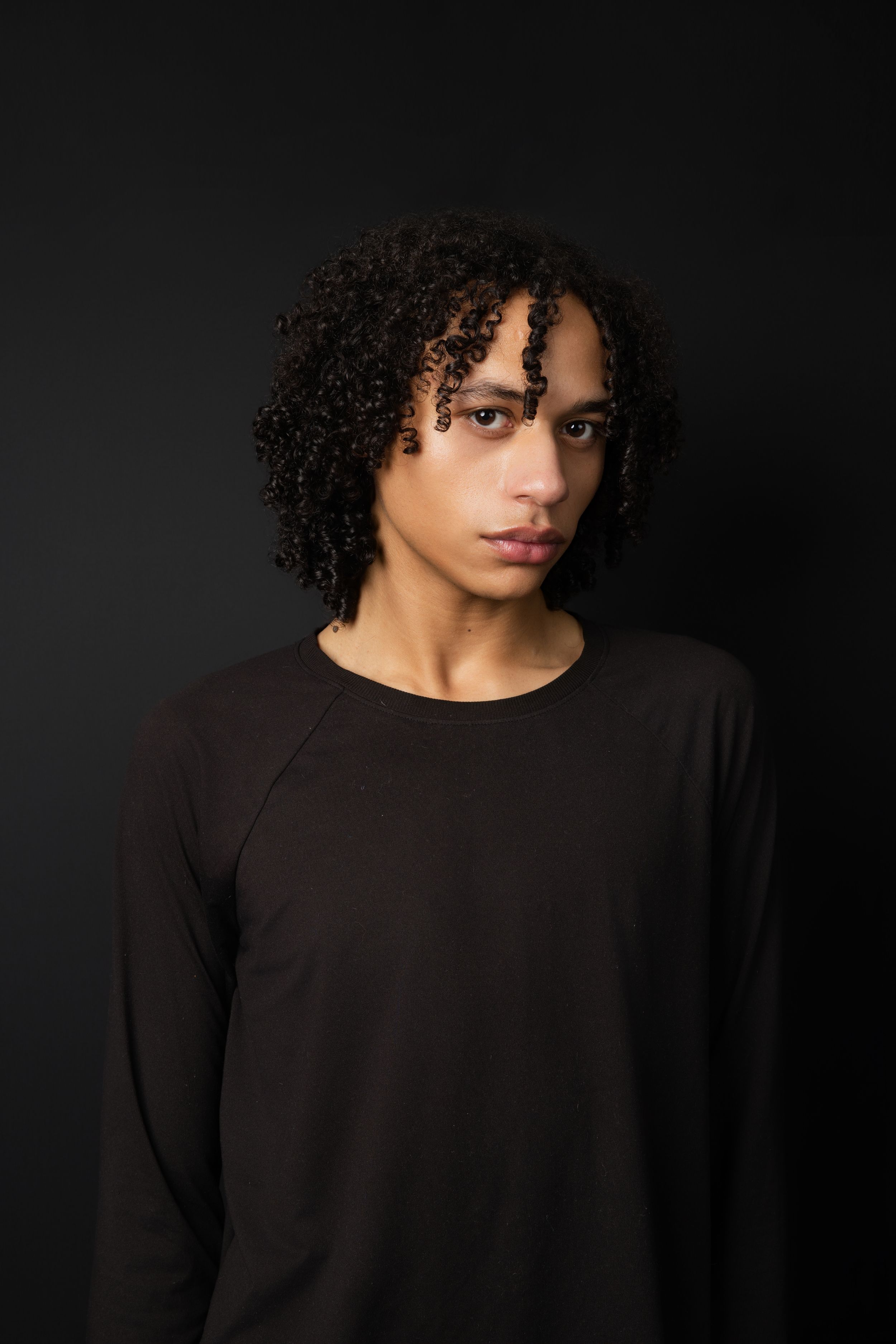 Young man with curly hair wearing a black long-sleeve shirt posed against a dark background.