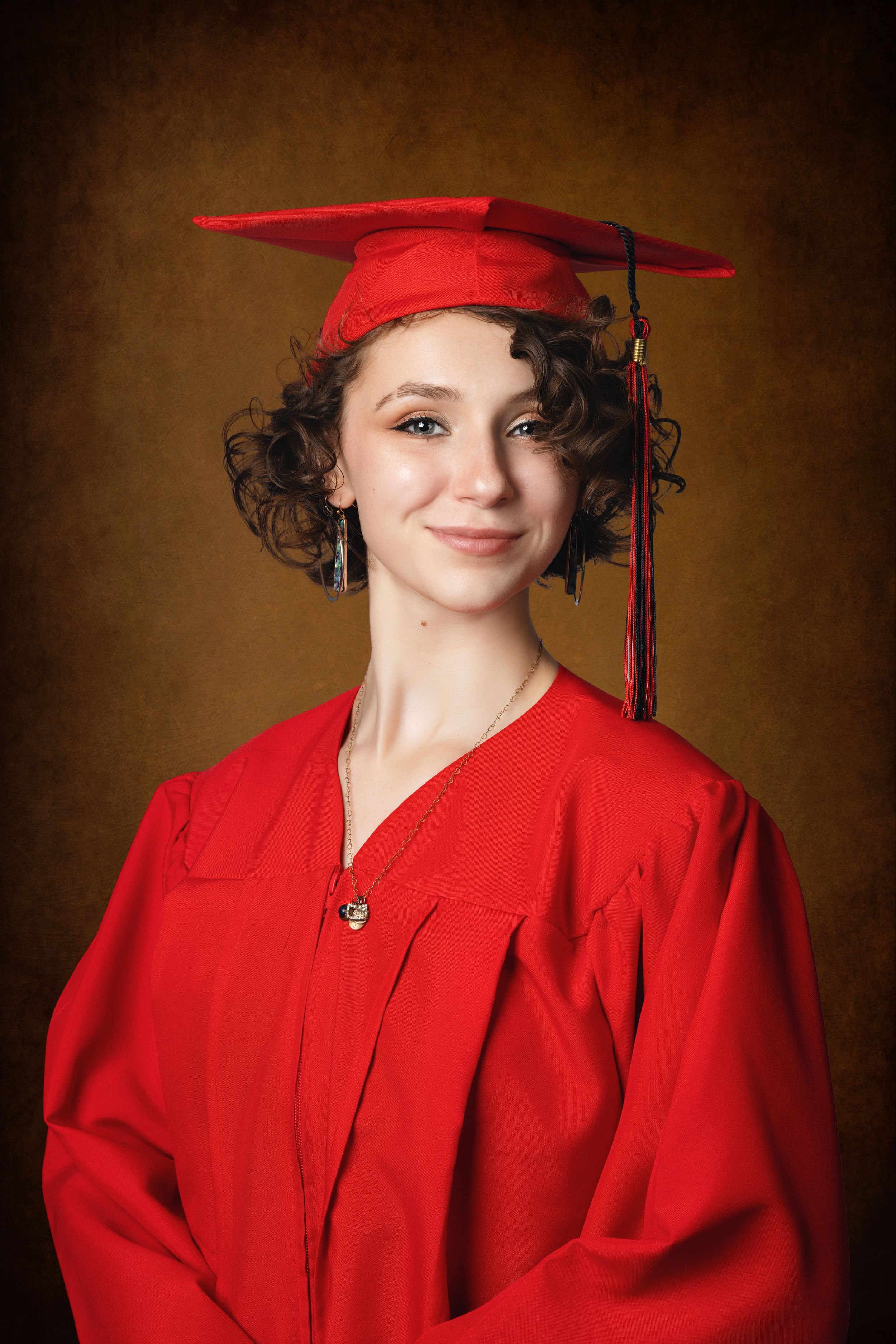 Young woman in red graduation cap and gown smiling for a portrait photo against a brown background.
