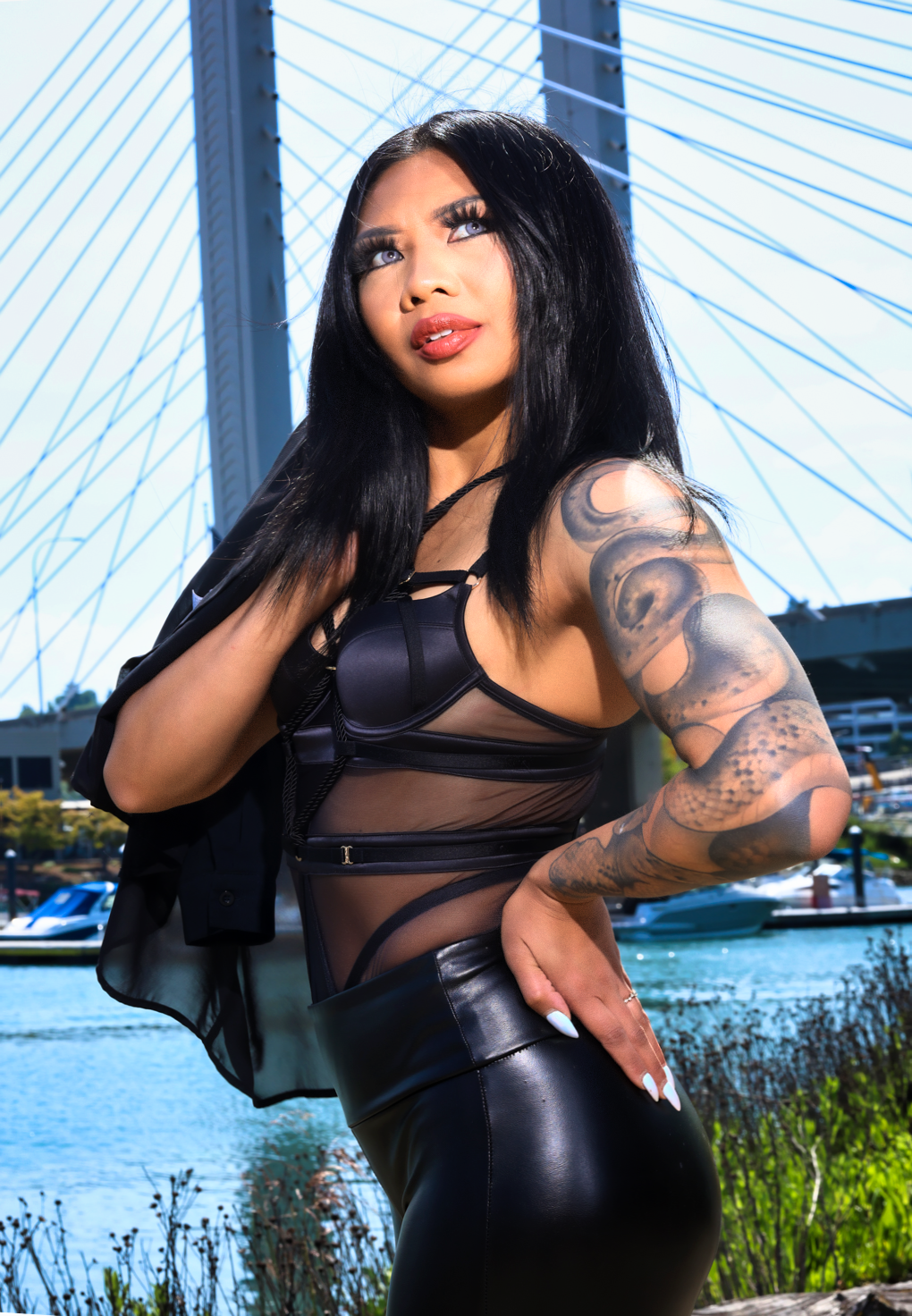 Woman with long black hair and sleeve tattoos poses in black mesh bodysuit and leather pants near a cable-stayed bridge.