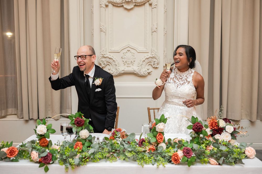 Bride and groom raising champagne glasses in toast at elegant wedding reception with floral garland centerpiece.