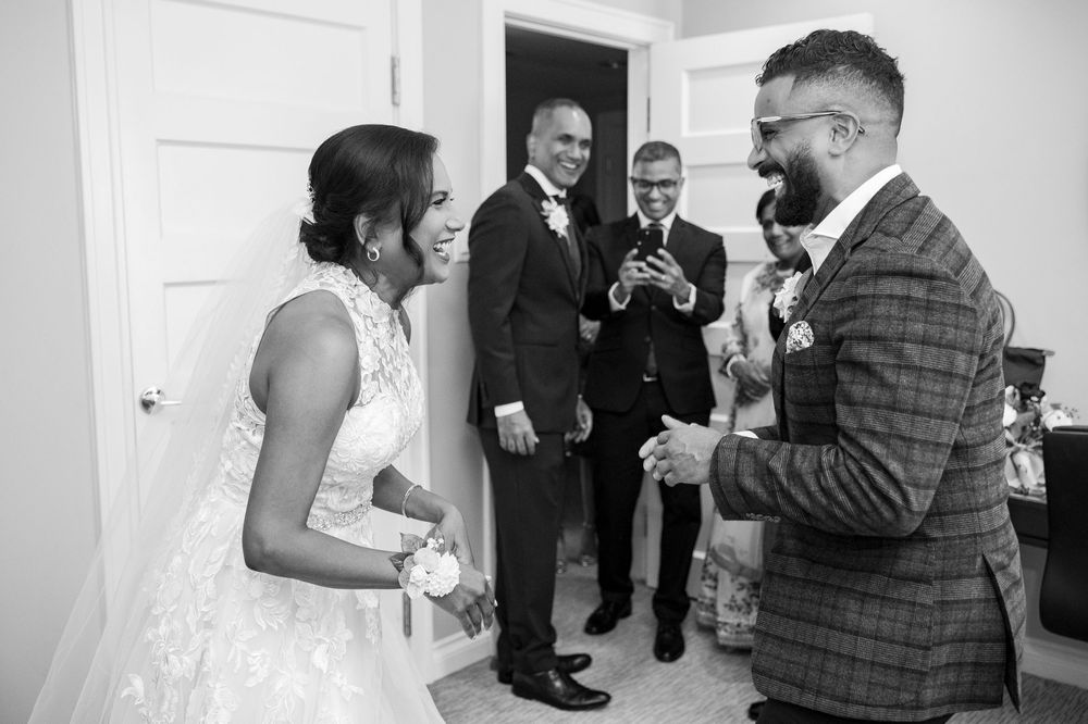 Black and white photo of a bride and groom sharing a joyful moment with guests during wedding preparations indoors.