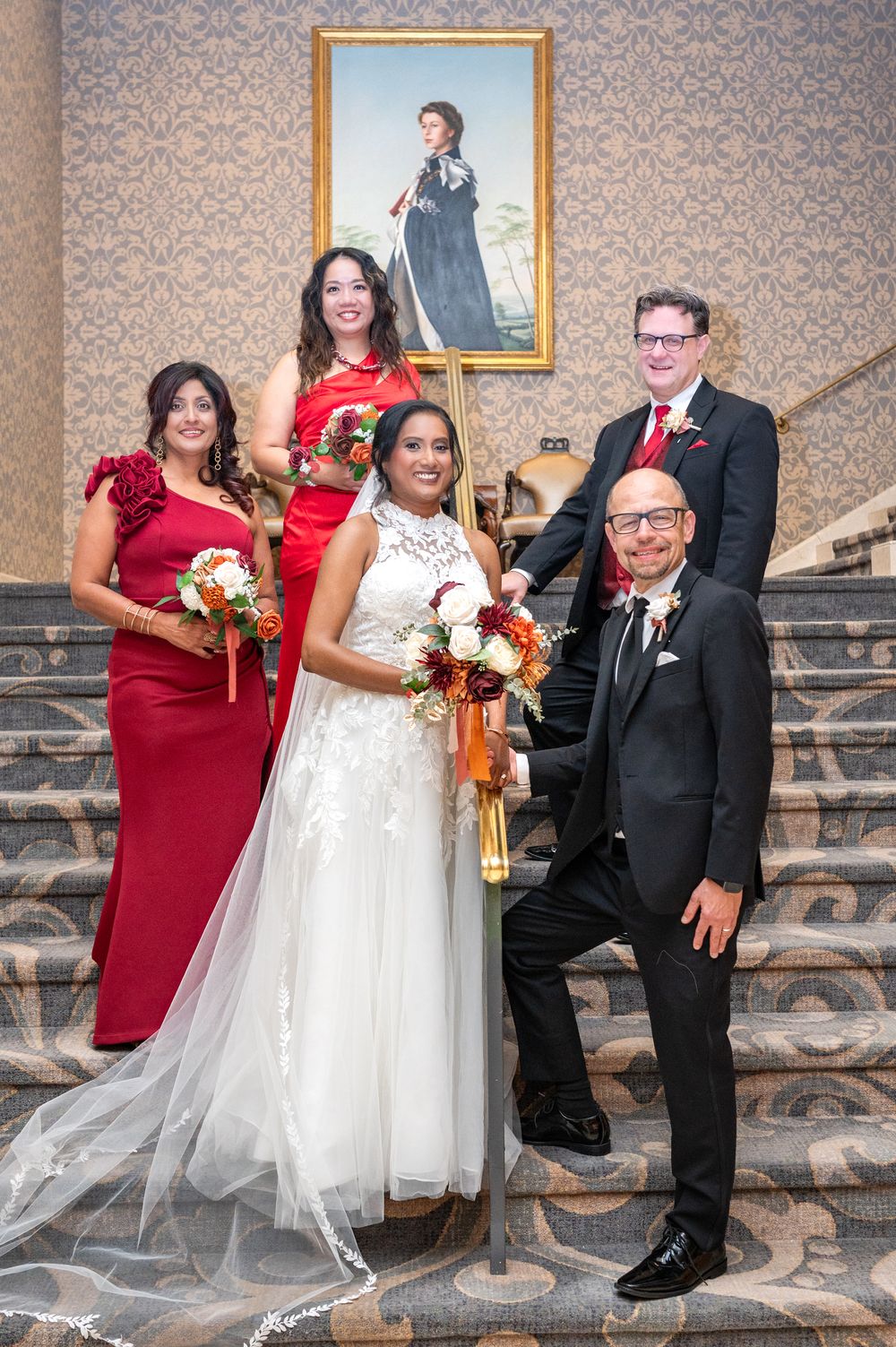 Wedding party poses on grand staircase, bride in white gown with bridesmaids in red dresses and groomsmen in black suits.