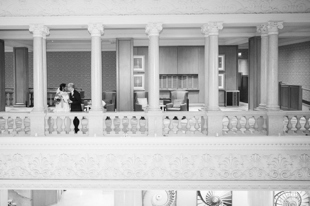 Black and white photo of a bride and groom sharing a romantic moment on an elegant balcony with ornate railings and columns.
