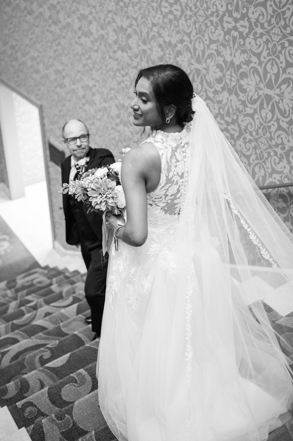 Bride in lace wedding gown holding bouquet with veil flowing, sharing a moment with older man in tuxedo.